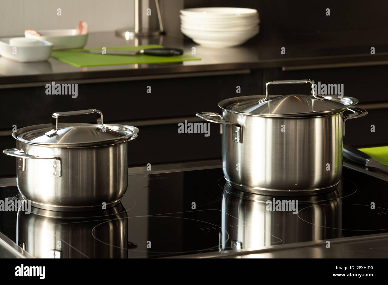 2 pots with lids are on a stove in the kitchen and are reflected in them. In the background are small plates, bowls and a knife. Stock Photo
