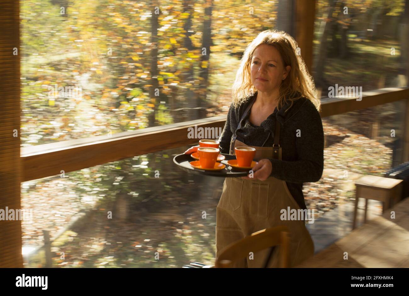 Female barista carrying coffee on tray along sunny autumn cafe window Stock Photo