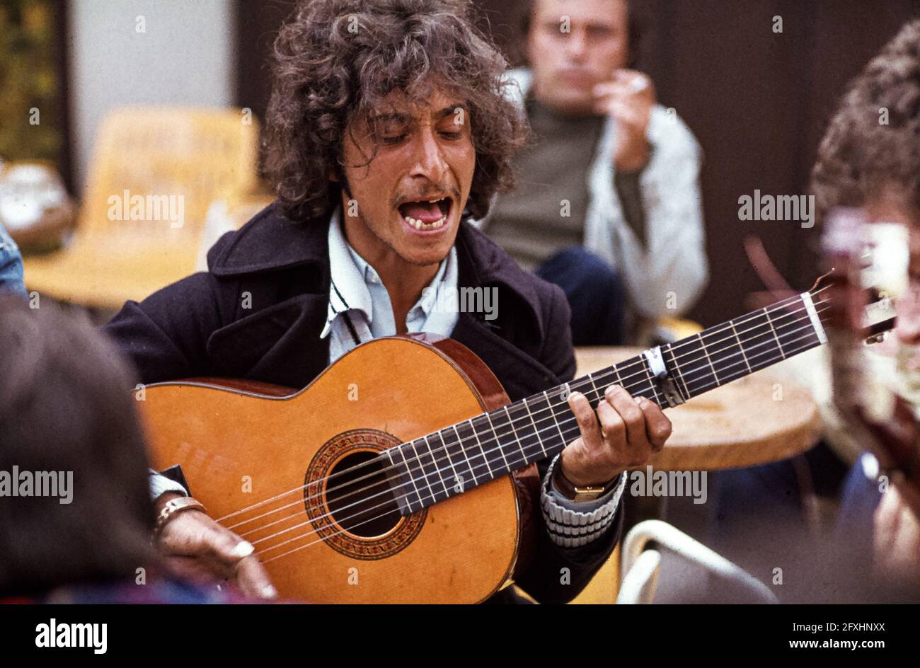 The guitarist sings with his eyes closed and his mouth wide open at a flamenco concert on the café terrace in Saintes-Maries-de-la-Mer; a member of the audience in the background is smoking. Roma pilgrimage, May 1978. Saintes-Maries-de-la-Mer, Arles, Bouches-du-Rhône, Provence-Alpes-Côte d'Azur, 13460, France Stock Photo