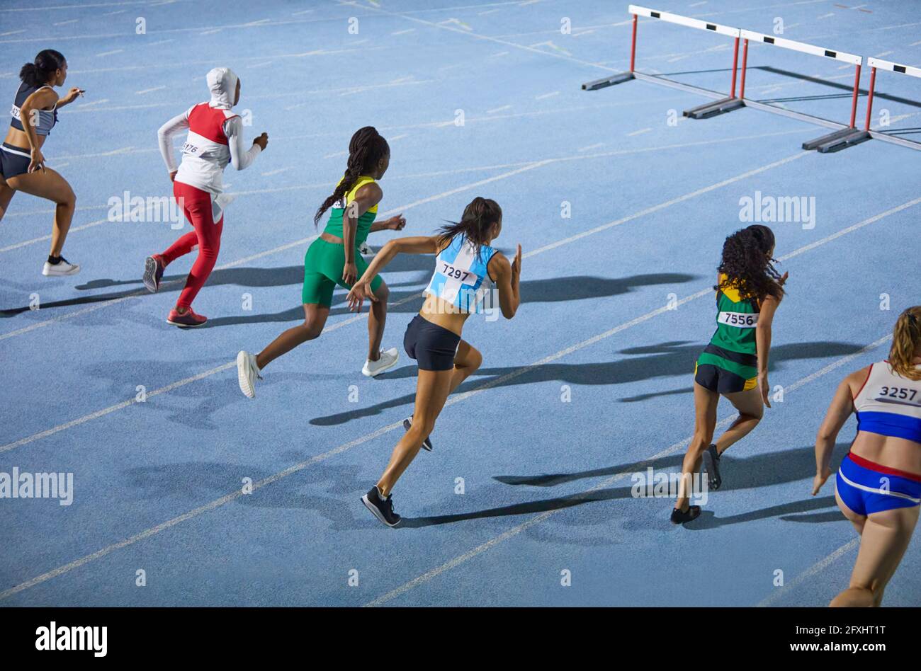 Female track and field athletes running hurdle race on blue track Stock