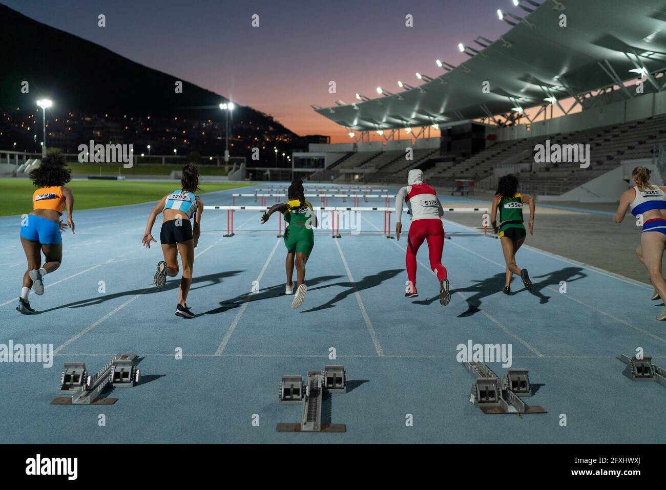 Female track and field athletes racing to hurdles on track at night