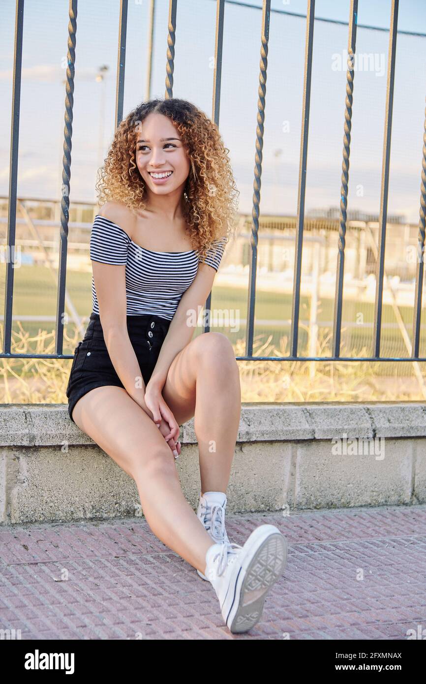 Young curly haired latina thought sitting on a fence. Cute latina girl in black shorts and striped t-shirt. High quality photo Stock Photo