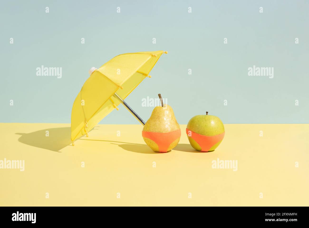 Summer fruit concept. Wet apple and a pear in monokini next to an umbrella isolated on a blue and yellow background. Abstract. Rectangle layout with c Stock Photo