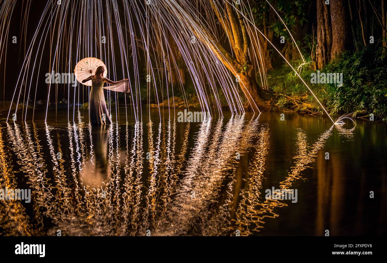 A young woman in a white dress standing in a river at night extends her hand to catch a falling spark, Kapaa, Hawaii, USA, Pacific Stock Photo