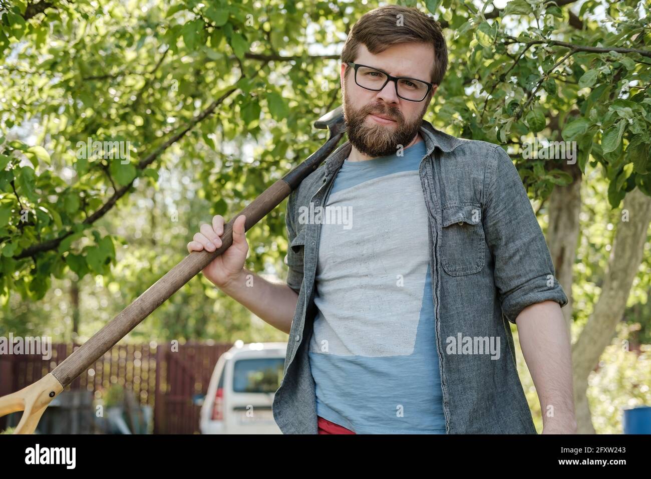 Tired man holds a shovel on his shoulder and looks at the camera, in the garden, against the background of a car and a fence. Stock Photo