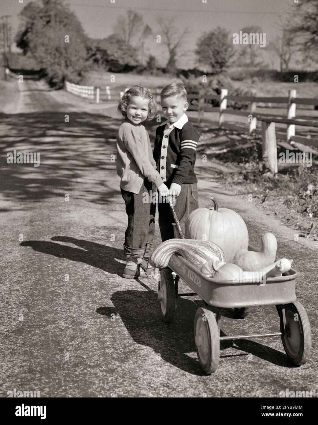 1950s TWO SMILING YOUNG KIDS BOY GIRL BROTHER SISTER IN BLUE JEANS LOOKING AT CAMERA PULLING WAGON FULL OF PUMPKINS AND GOURDS - c4928 HAR001 HARS SISTER 1 WAGON JUVENILE CUTE STYLE FRIEND TEAMWORK JOY LIFESTYLE FEMALES BROTHERS YOUTHS RURAL HEALTHINESS HOME LIFE NATURE TRANSPORT FRIENDSHIP FULL-LENGTH HALF-LENGTH PERSONS MALES SIBLINGS SISTERS TRANSPORTATION AGRICULTURE B&W EYE CONTACT HARVESTING HAPPINESS PUMPKINS AND FALL SEASON SIBLING GOURD FRIENDLY YOUNGSTERS VARSITY SWEATER OCTOBER CHARMING COOPERATION GOURDS GROWTH JUVENILES SEASON TOGETHERNESS YOUNGSTER AUTUMNAL BLACK AND WHITE Stock Photo