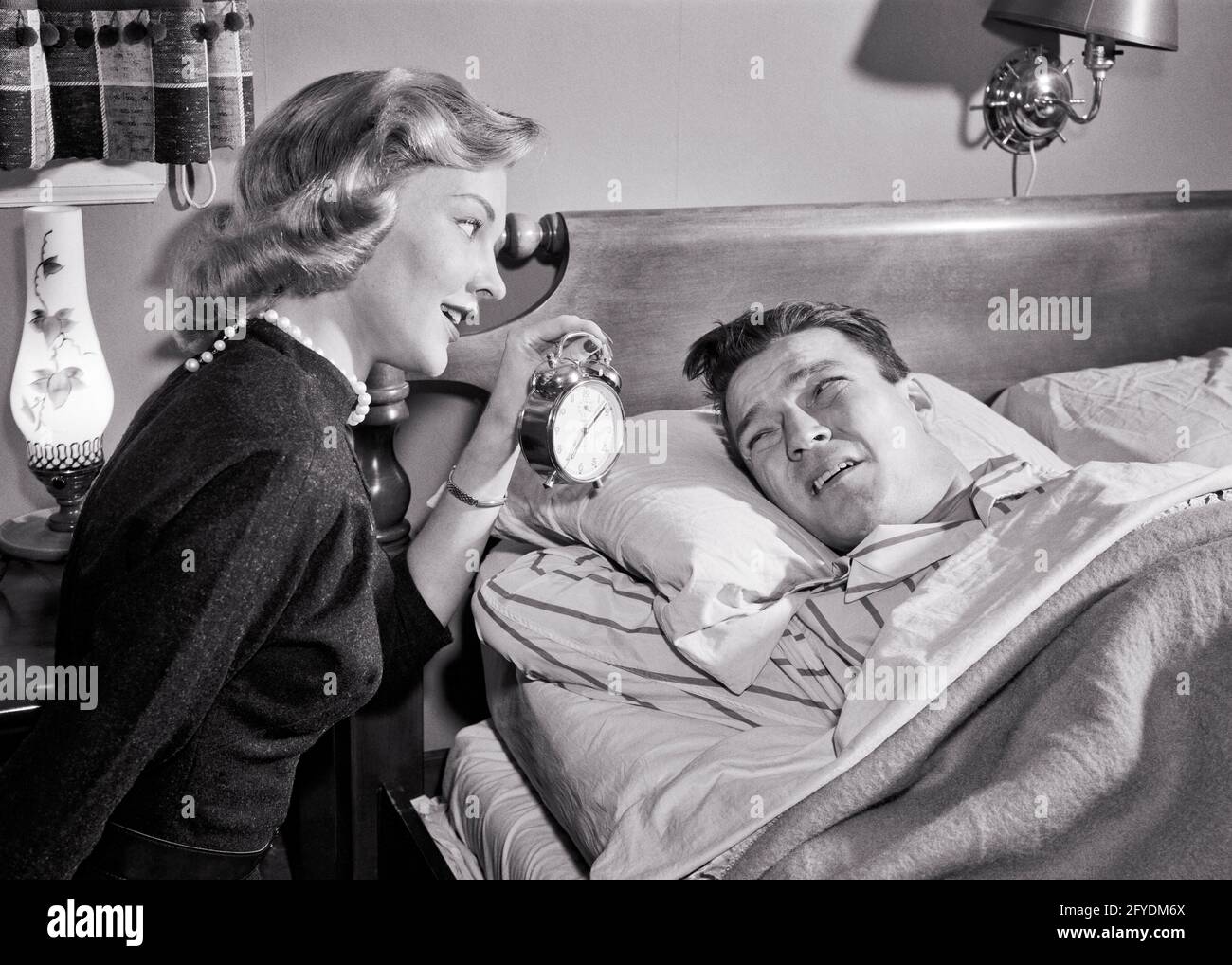 1950s CHEERFUL BLONDE WIFE HOLDING UP ALARM CLOCK TO HER SLEEPING WAKING  HUSBAND STILL IN BED AT 7.08 AM WORKDAY SCHEDULE - s8303 DEB001 HARS 1  COMMUNICATION PEACE JOY LIFESTYLE ANNOYED FEMALES