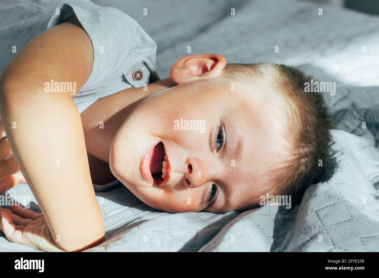 Portrait 4 or 5 years old little cute smiling and having fun boy on a bed in bedroom. A brunette boy in a grey shirt. Stock Photo