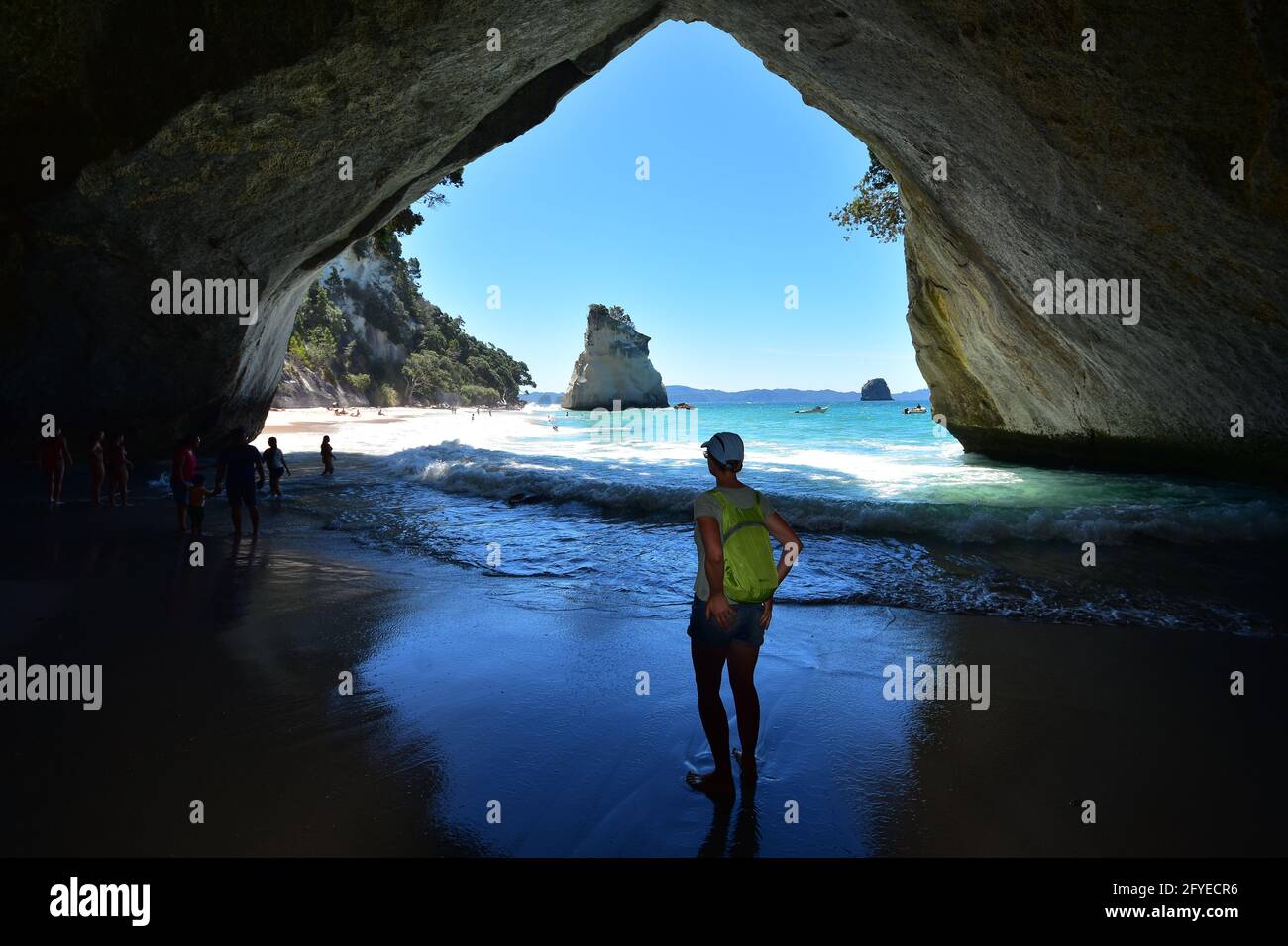 Female tourist with green backpack and other holidaymakers under natural rock arch in Cathedral Cove. Stock Photo