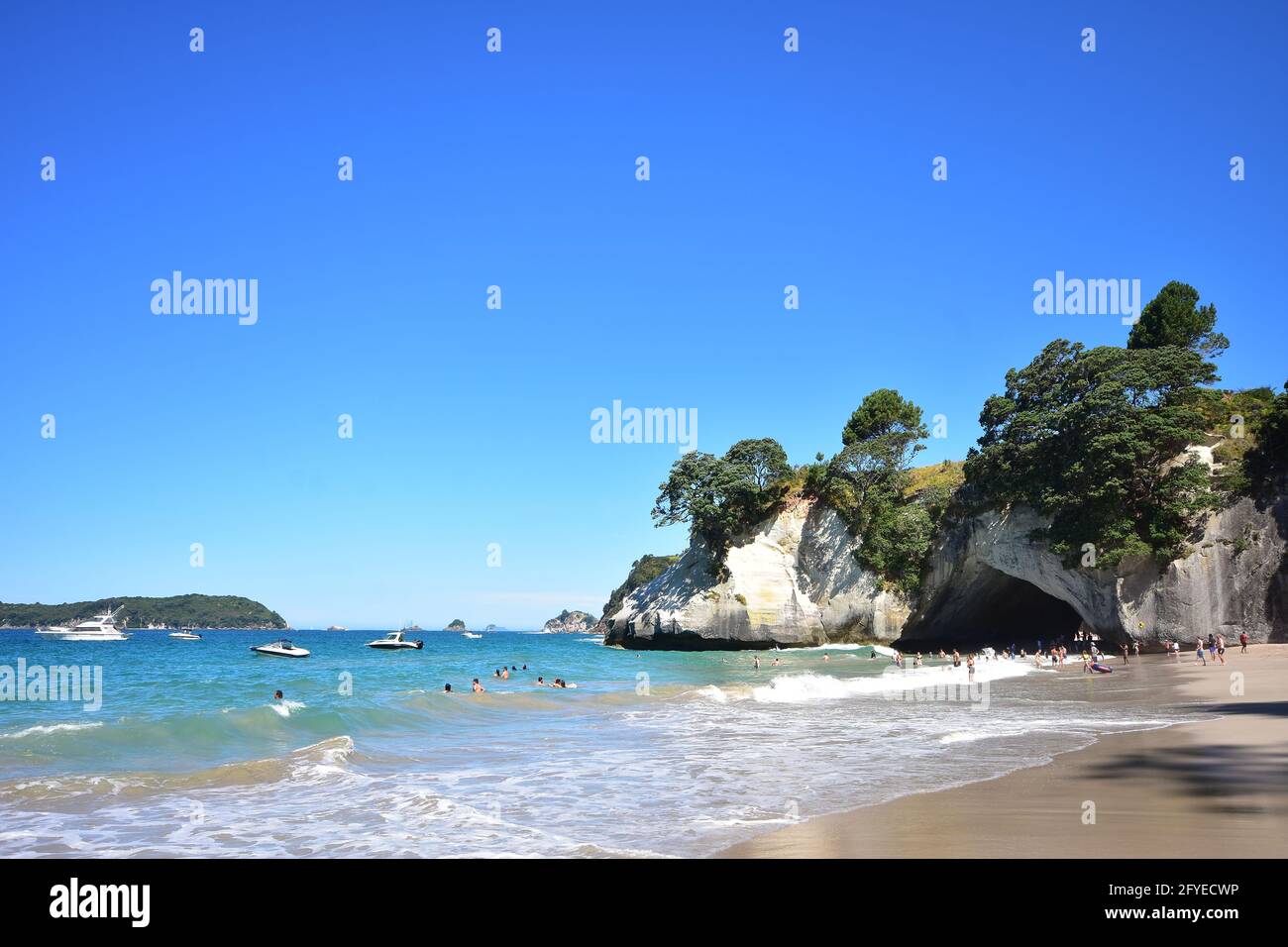Holidaymakers playing and swimming in surf waves coming to sandy beach on Cathedral Cove with anchored boats and distant islands in background. Stock Photo