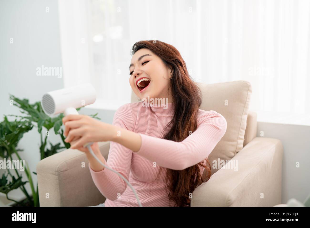Happy young woman blow drying hair in living room while feeling excited emotions Stock Photo - Alamy