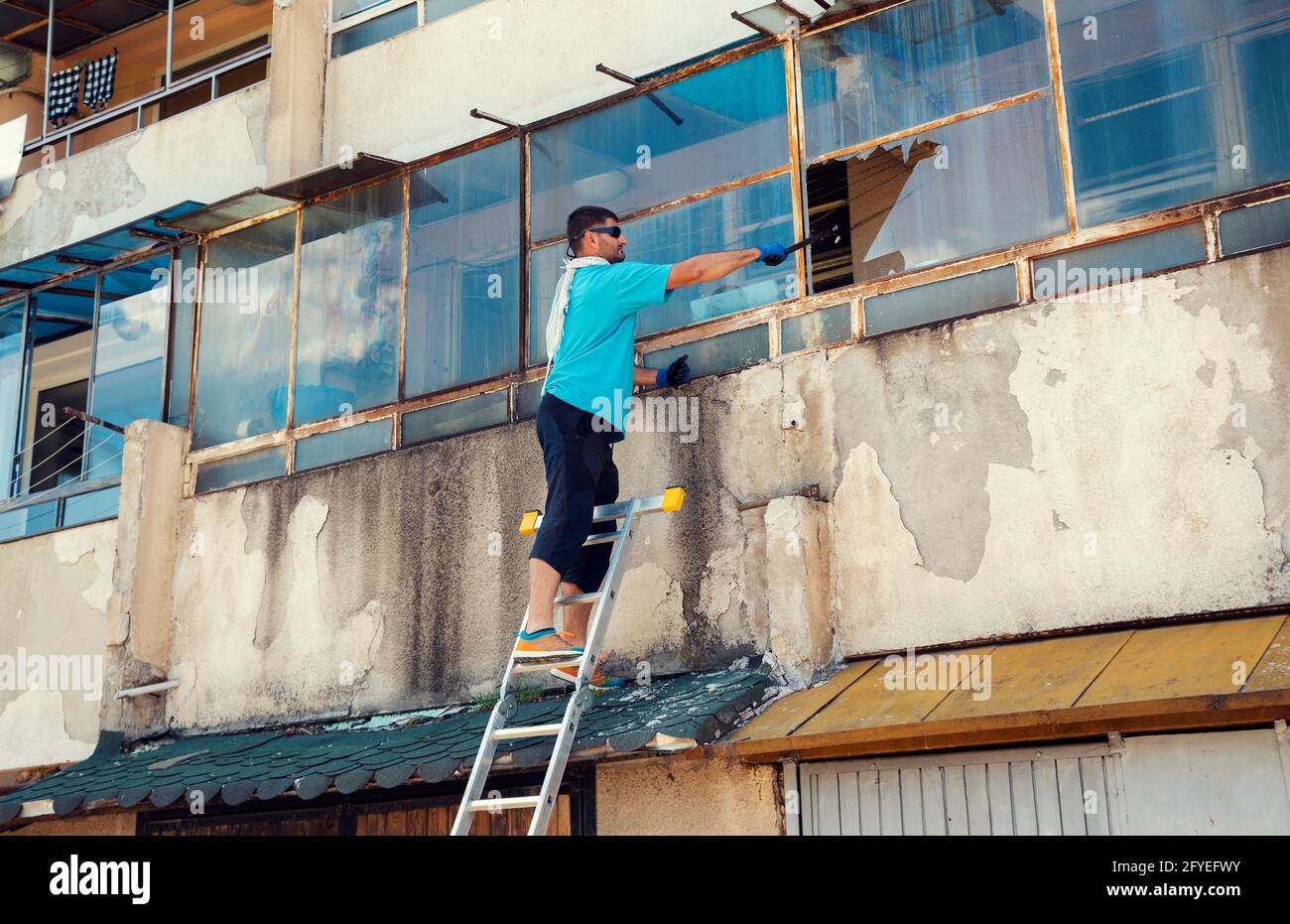 Caucasian man breaking a window to rescue his neighbor standing on a ladder and wearing a protective glove and sunglasses Stock Photo