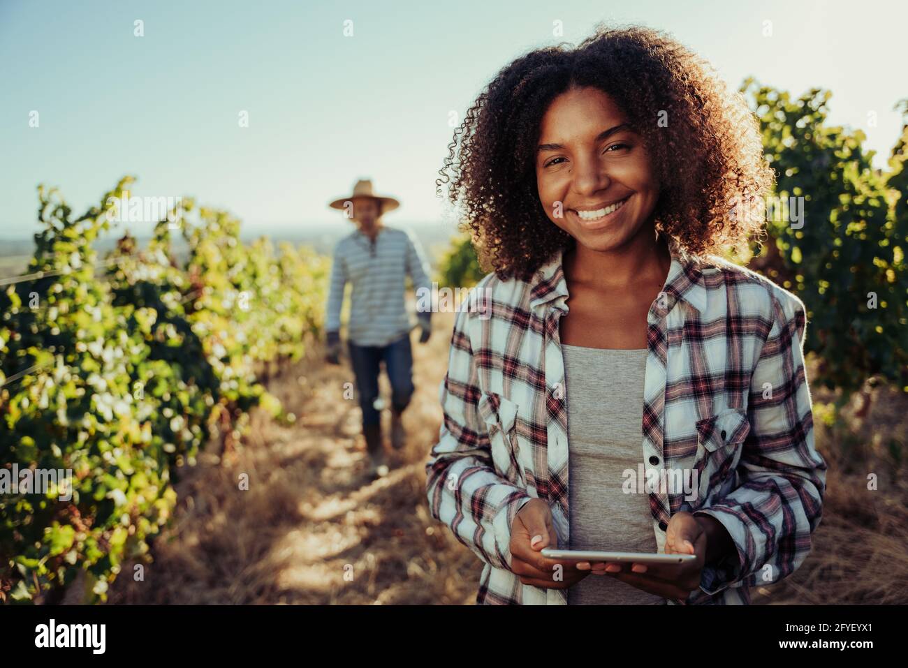 Female farmer standing in vineyards holding digital tablet while male colleague examines plants behind Stock Photo