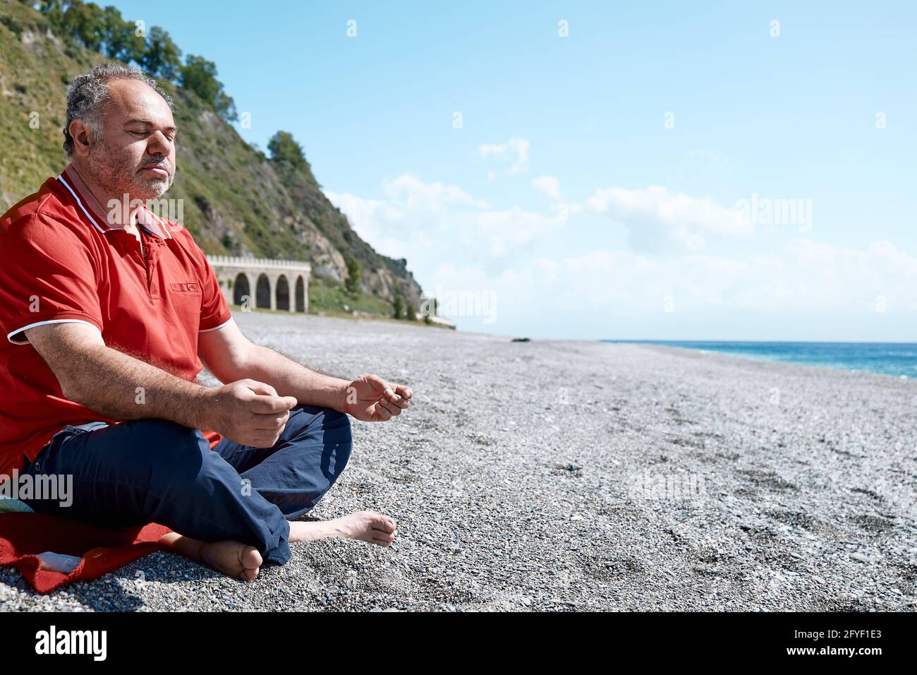 Full length of middle aged bearded man in casual clothes sitting in lotus pose on pebble beach and meditating on sunny day. Healthy lifestyle. Yoga Stock Photo