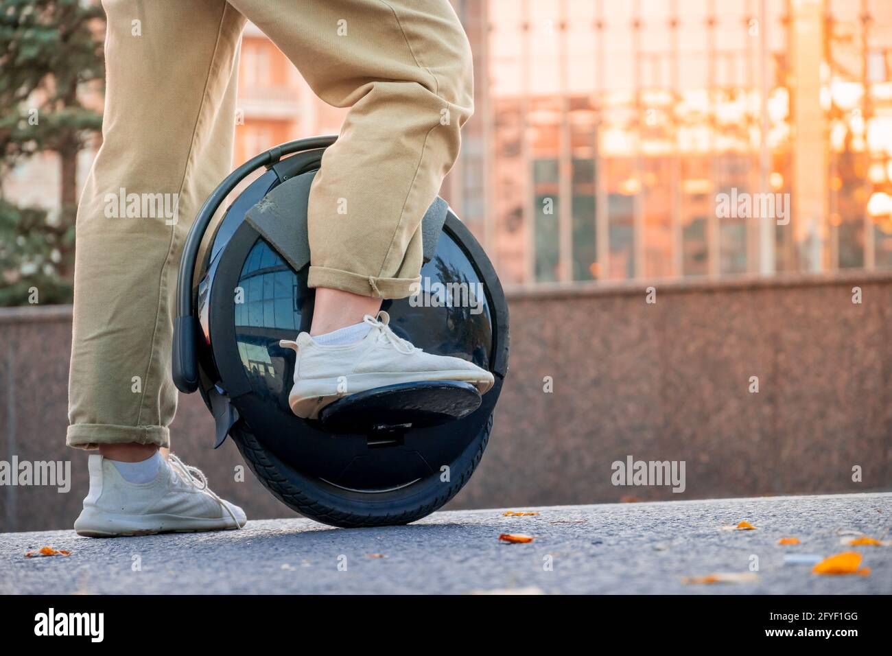 The legs of a young woman ready to ride an electric unicycle, abbreviated EUC, against the background of an evening or morning urban landscape. An unr Stock Photo