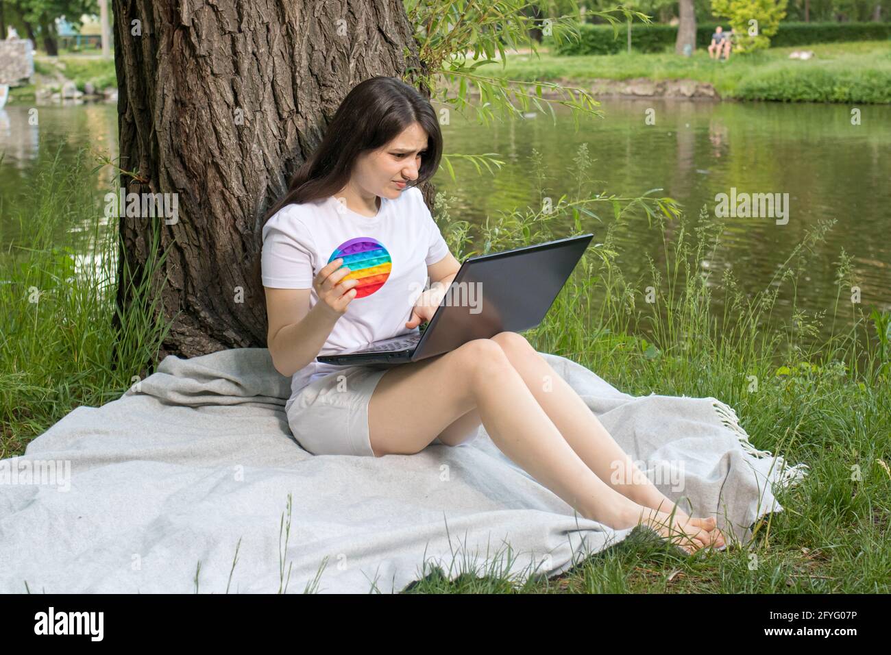 A woman freelancer works at a laptop in nature and pops it with a reusable bubble wrap. Coping with stress at work with Pop it Stock Photo