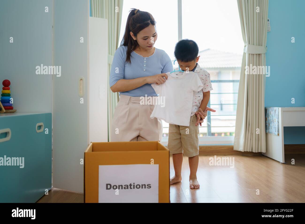 Asian mother and son are standing near closet of clothes in the dressing room carrying a box of clothes donated to take to the donation center. Stock Photo