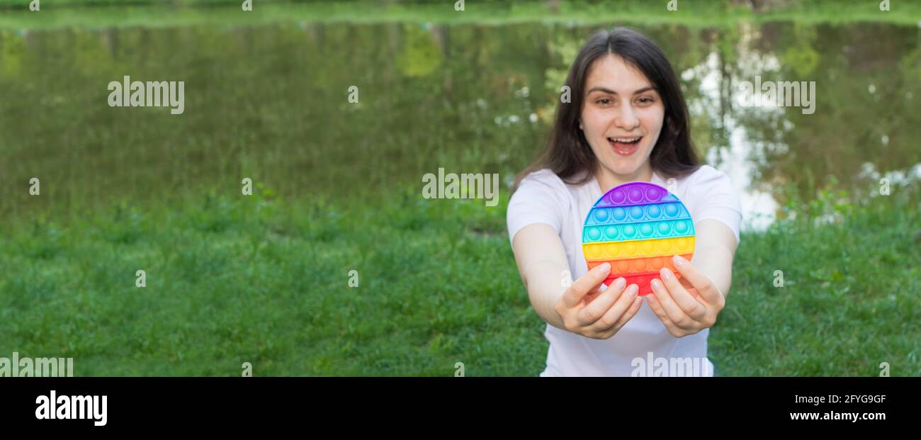 Brunette girl plays with a toy pop it on nature. Long horizontal banner. Trend 2021 is a reusable bubble wrap, a funashionable game against stress Stock Photo