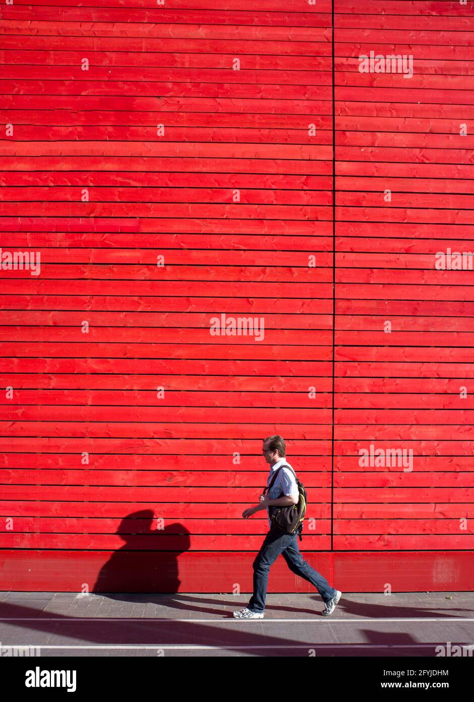 Sideview of a man, with a rucksack bag, walking past a red wooden wall with his shadow on it on a warm sunny day Stock Photo