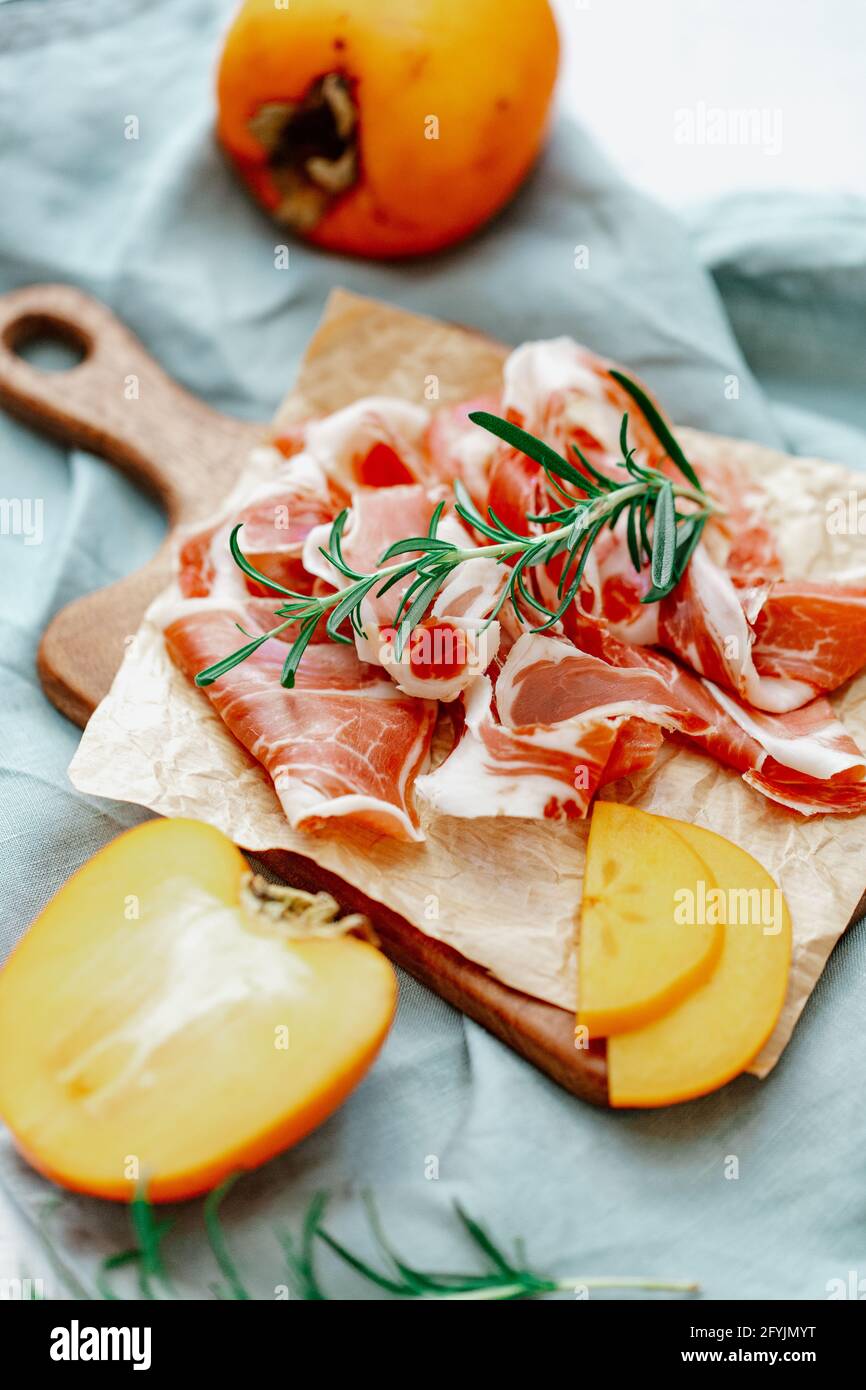 Slices of fresh prosciutto and persimmon fruit on a wooden chopping board Stock Photo