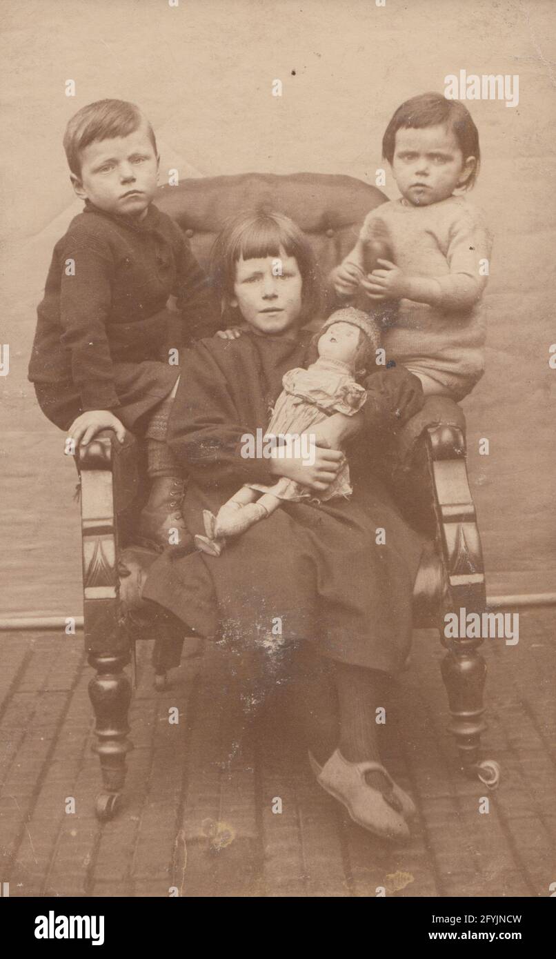 Vintage early 20th century photographic postcard showing three poor looking children sat on a chair. The older girl is holding a doll. Stock Photo