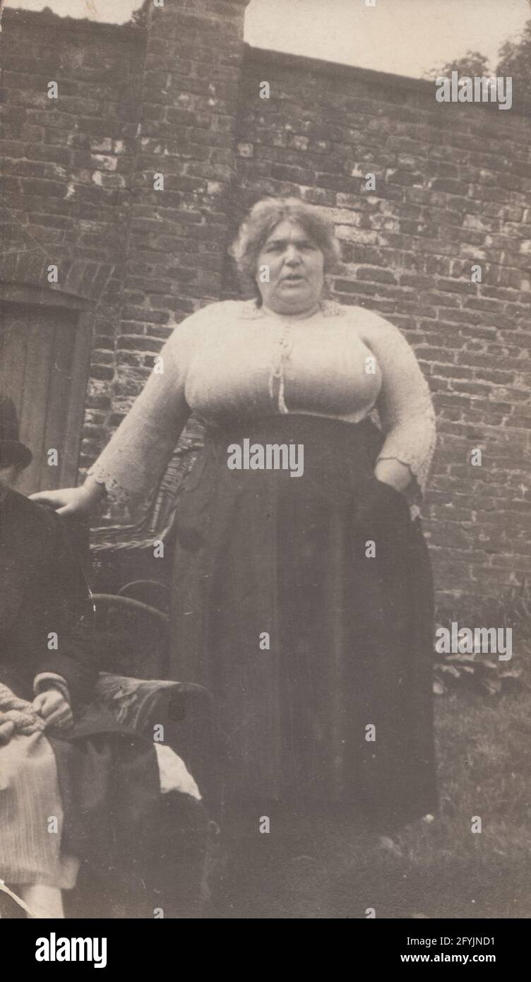 Vintage early 20th century photographic postcard showing a buxom lady stood in a garden. Stock Photo