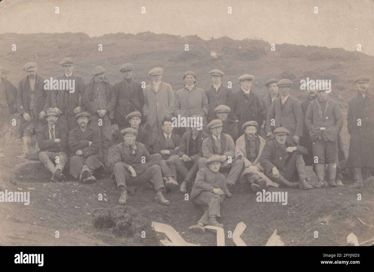 Vintage early 20th century photographic postcard showing a large group of working men wearing caps in a remote location. Stock Photo