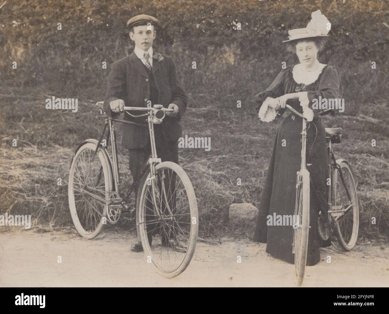Vintage British postcard showing a smartly dressed man and a woman stood outdoors holding their pedal cycles. Stock Photo