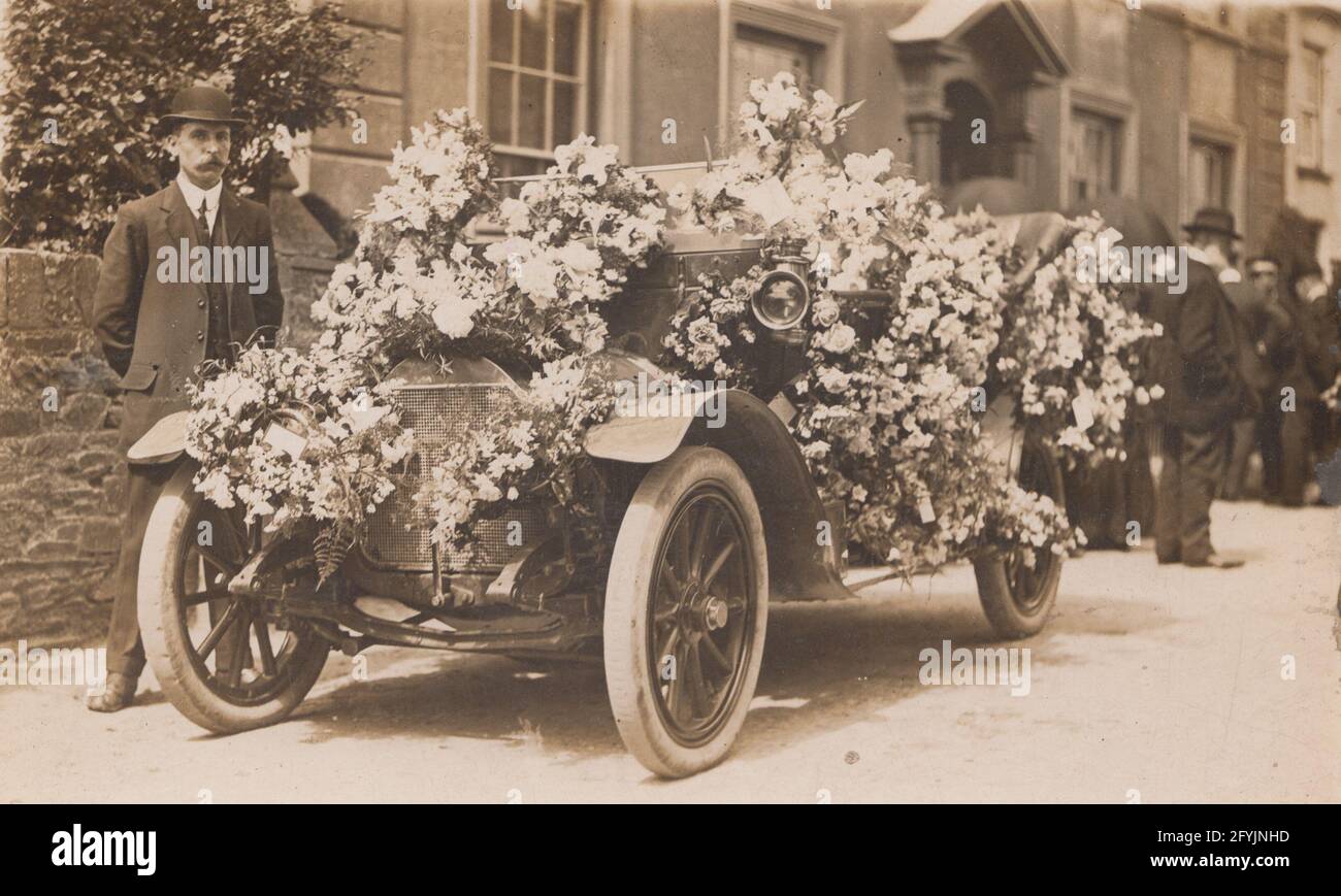 Vintage early 20th century photographic postcard showing a man standing next to an automobile covered in flowers. Dressed up for a wedding or funeral. Stock Photo
