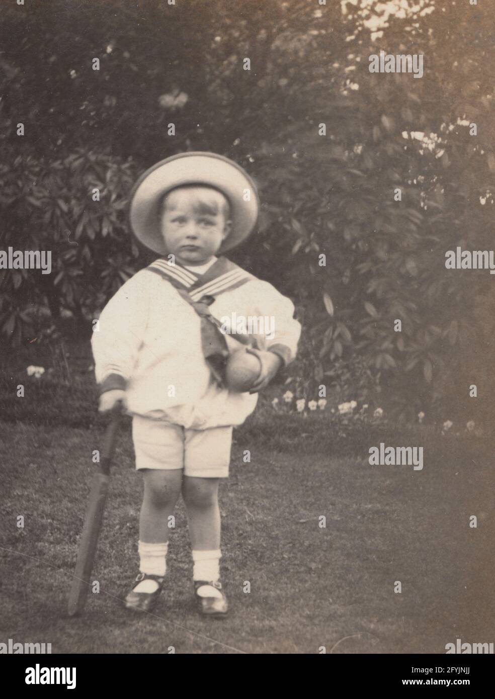 Vintage early 20th century photographic postcard showing a young child stood in a garden wearing a sailors outfit. Holding a cricket bat and ball. Stock Photo