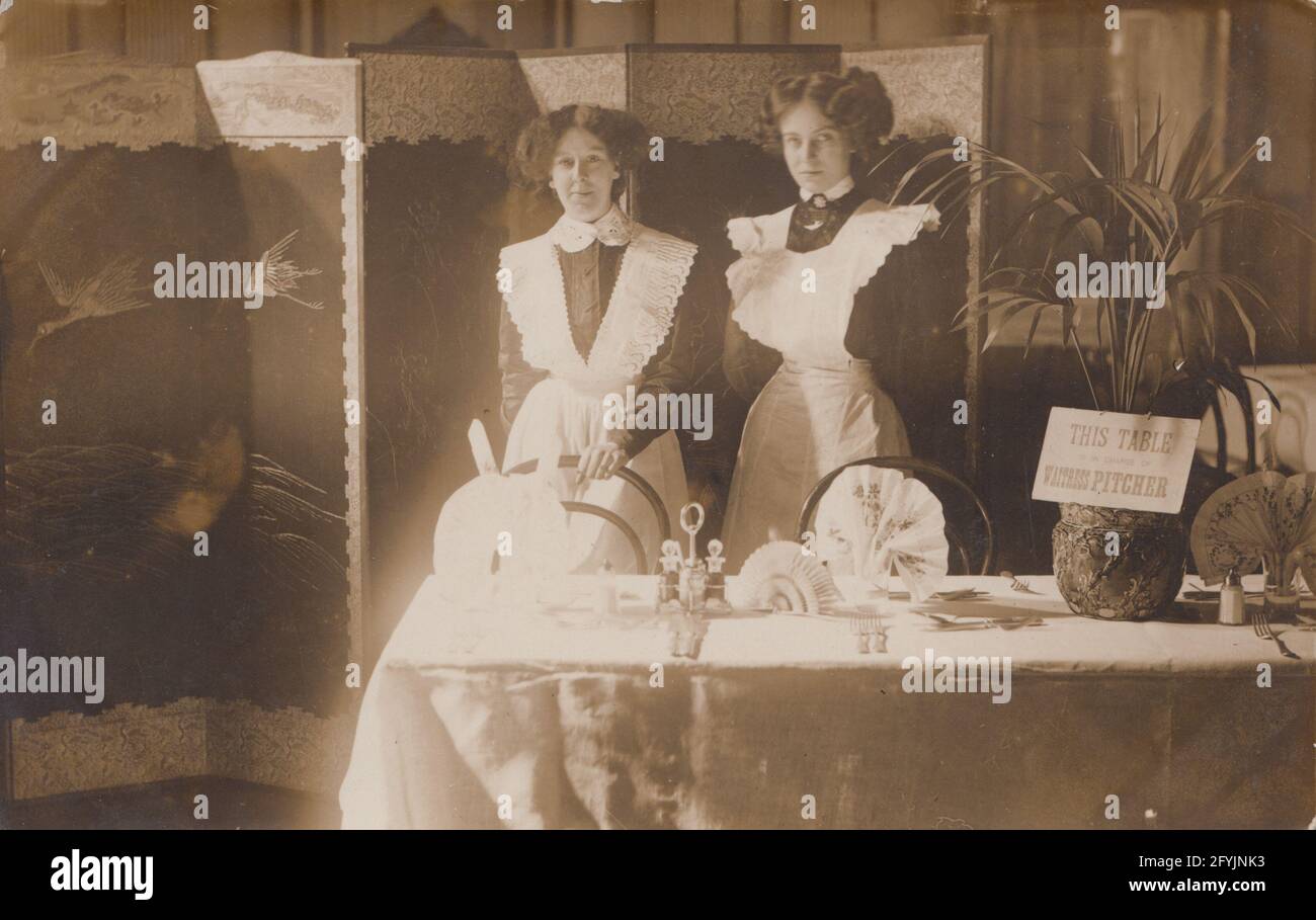 Vintage early 20th century photographic postcard showing two British waitresses stood at the dinner table. Stock Photo