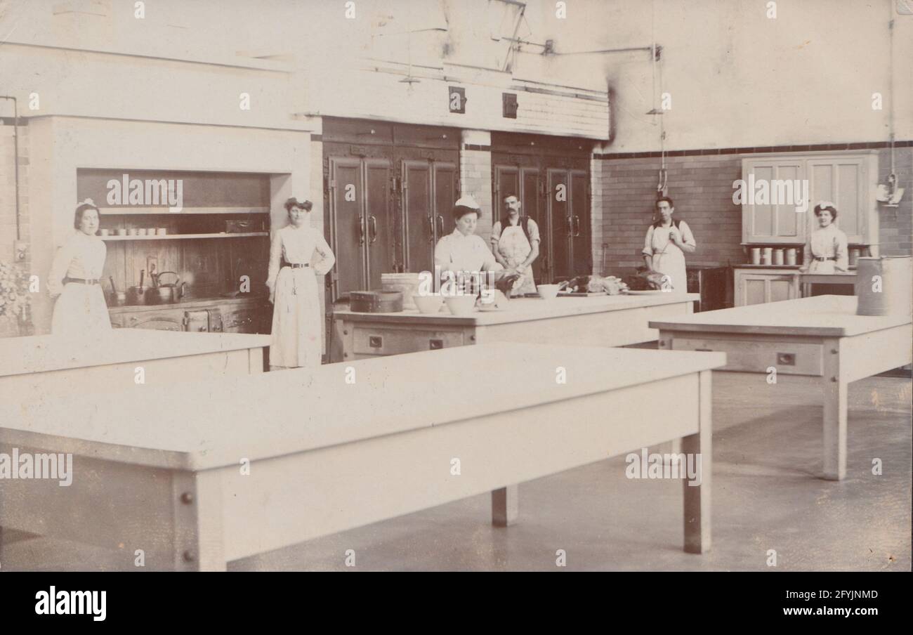 Vintage Early 20th Century Photographic Postcard Showing a group of staff in the kitchen of a stately home. Lady preparing food at one of the tables. Stock Photo