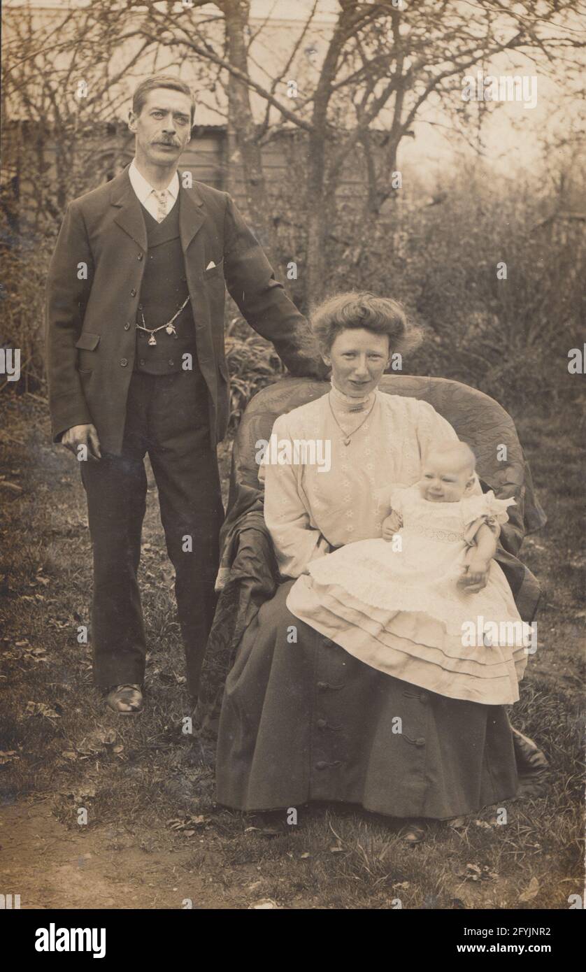 Vintage early 20th century photographic postcard showing a mother and father posing in a garden with their baby. Stock Photo