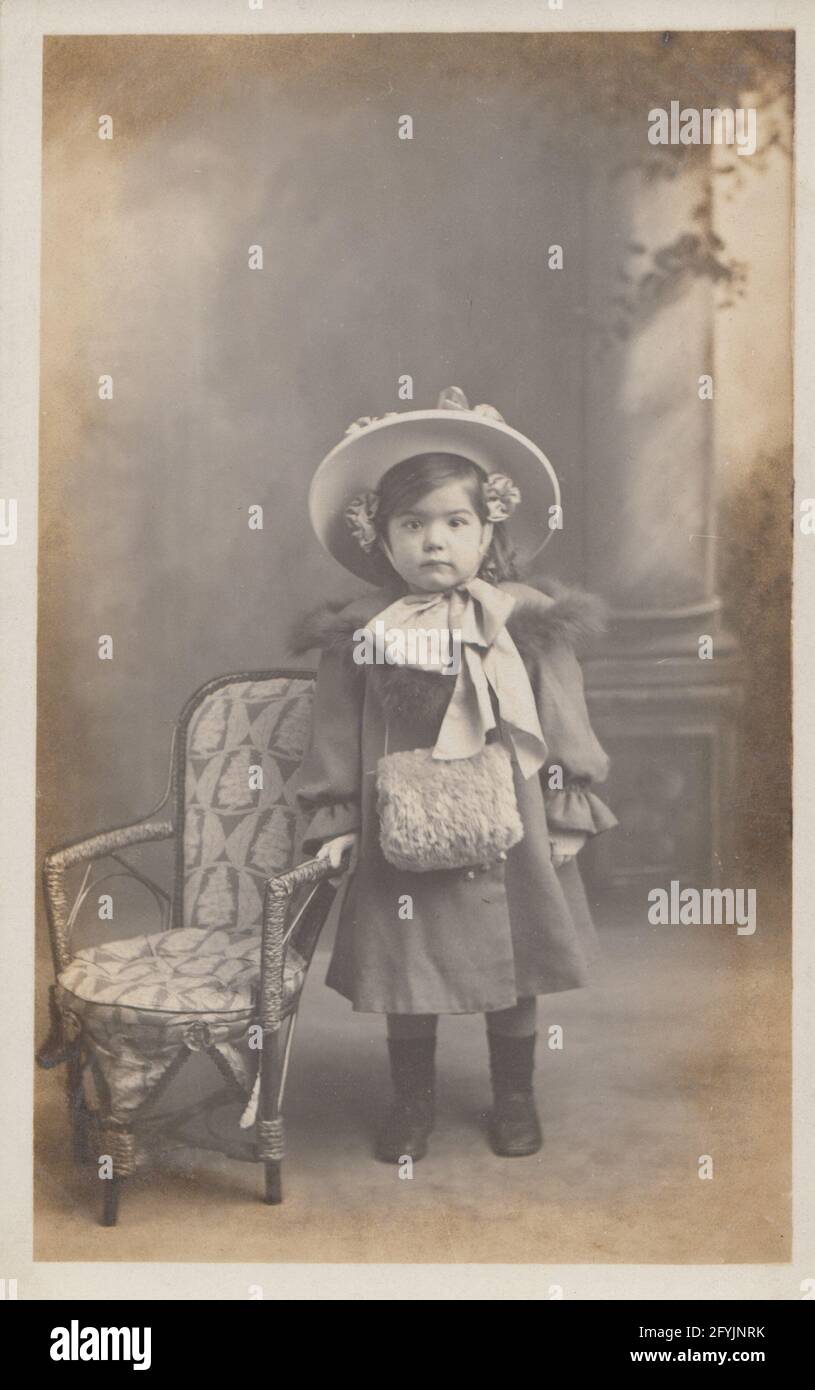 Vintage early 20th century photographic postcard showing a beautiful young girl in her coat and bonnet. Stock Photo