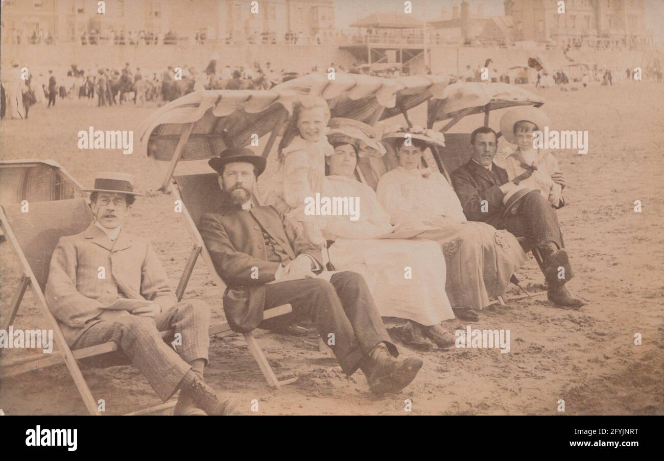 Vintage early 20th century photographic postcard showing a family group sitting in deck chairs and enjoying themselves at the seaside. Stock Photo