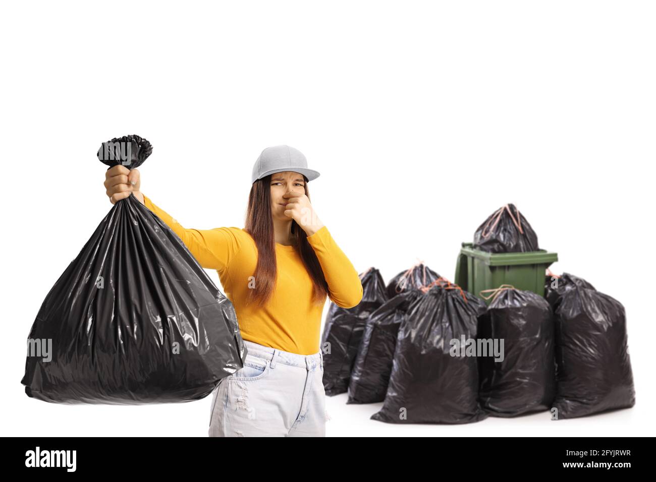 Young female holding a smelly bin bag in front of a bin and a pile of black bags isolated on white background Stock Photo