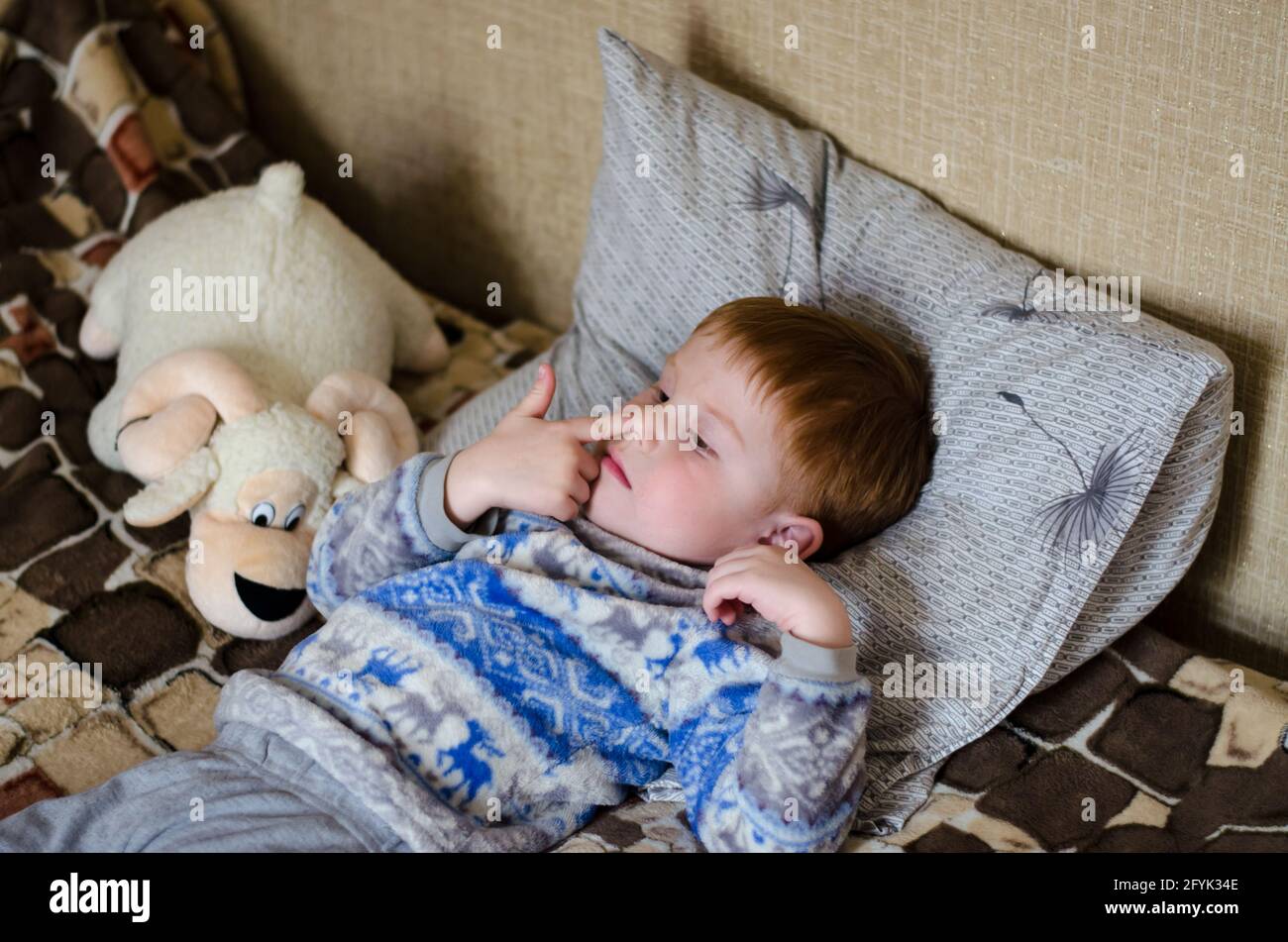 A 4-year-old red-haired boy is lying on the bed and picking his nose with his finger. Child picking his nose Stock Photo