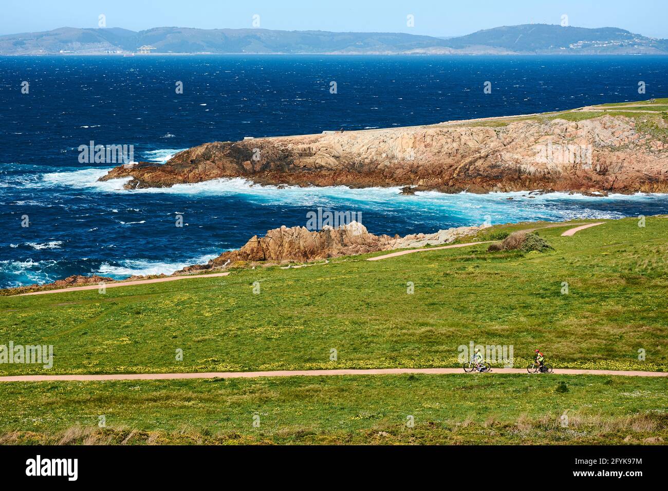 Two cyclists riding their bikes on path near the Tower of Hercules in Coruña, Spain, surrounded by green grass and flowers with view of the coastline Stock Photo