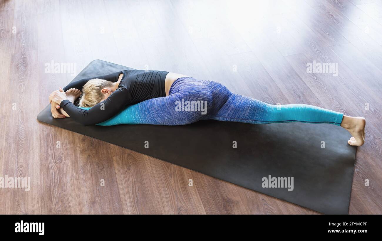 A woman in sportswear practicing yoga performs hanumanasana exercise on a mat in the studio, a longitudinal split with the head tilted to the leg. Stock Photo