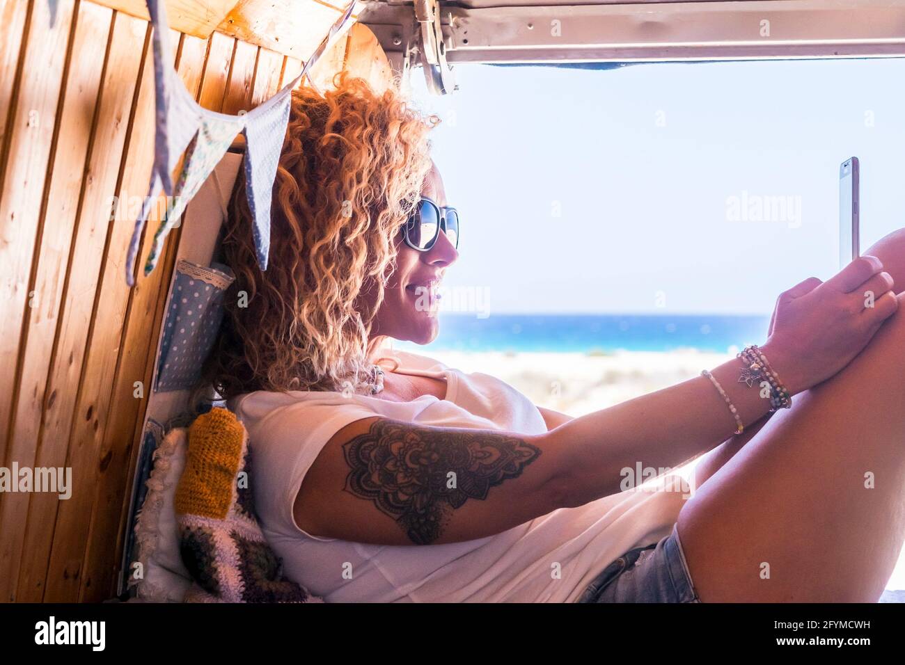 Happy adult woman use mobile phone connection sitting and relaxing inside old vintage wooden van in summer travel holiday vacation lifestyle alone - b Stock Photo