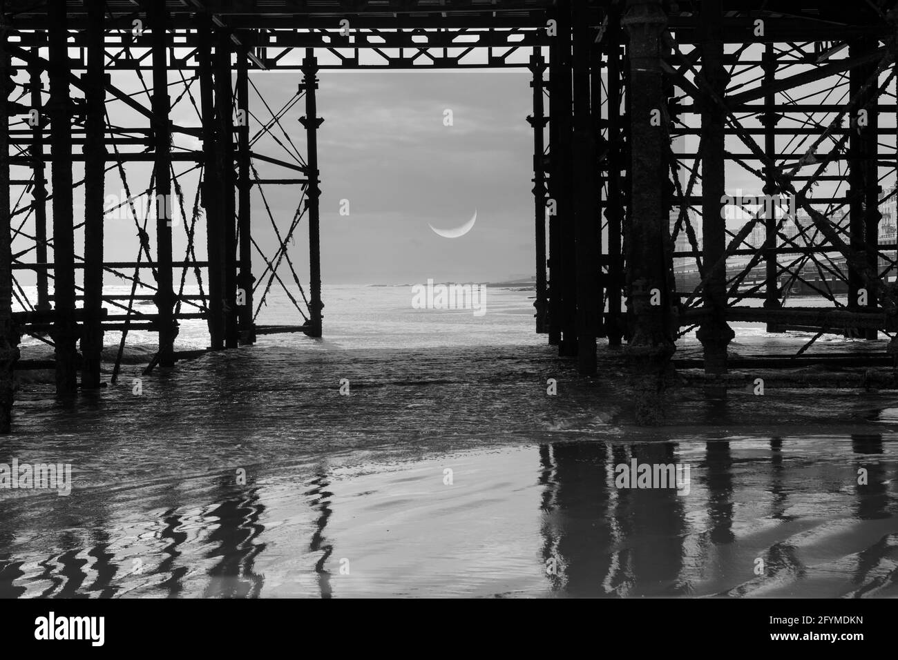 Crescent moon viewed through a pier with reflections in the wet sand Stock Photo