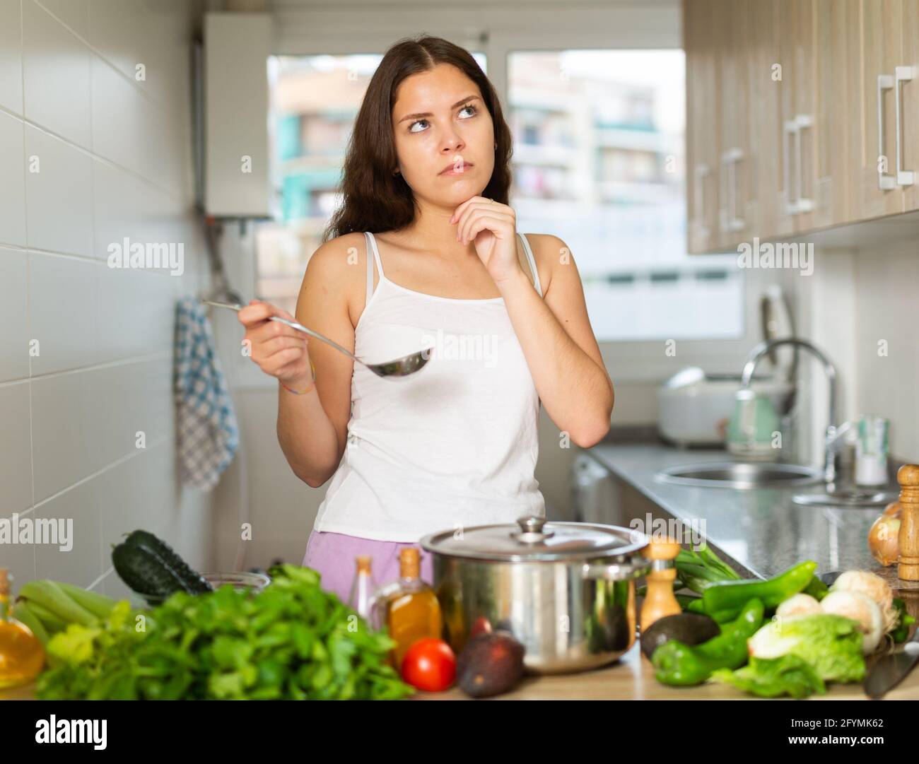 Pensive young brunette woman standing at kitchen thinking about cooking ...