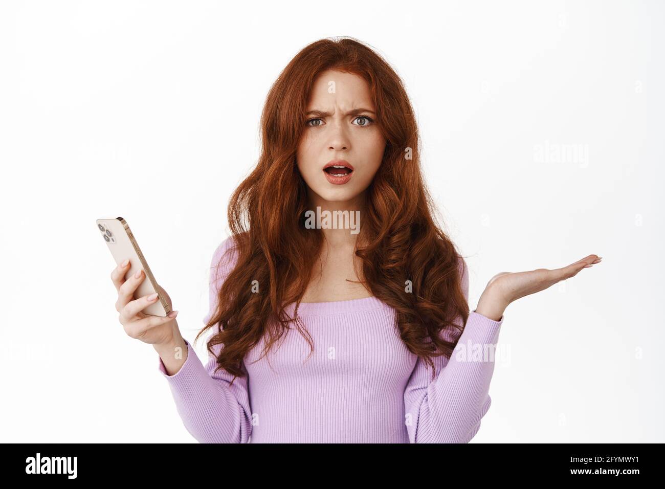 Confused ginger girl shrugging shoulders with smartphone, looking questioned at camera, being frustrated and annoyed by something on mobile phone Stock Photo
