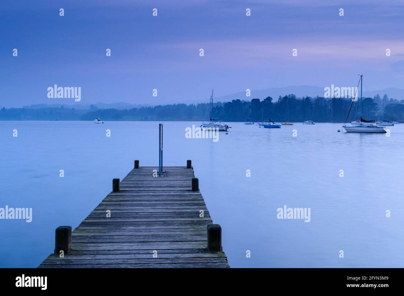 View from small wooden jetty over Windermere (boats on calm water, boardwalk, dramatic blue hour evening light) - Lake District, Cumbria, England UK. Stock Photo