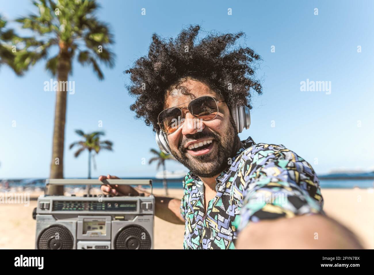 Young Latin man having fun taking selfie with mobile smartphone while listening music with headphones and boombox on the beach during summer vacations Stock Photo
