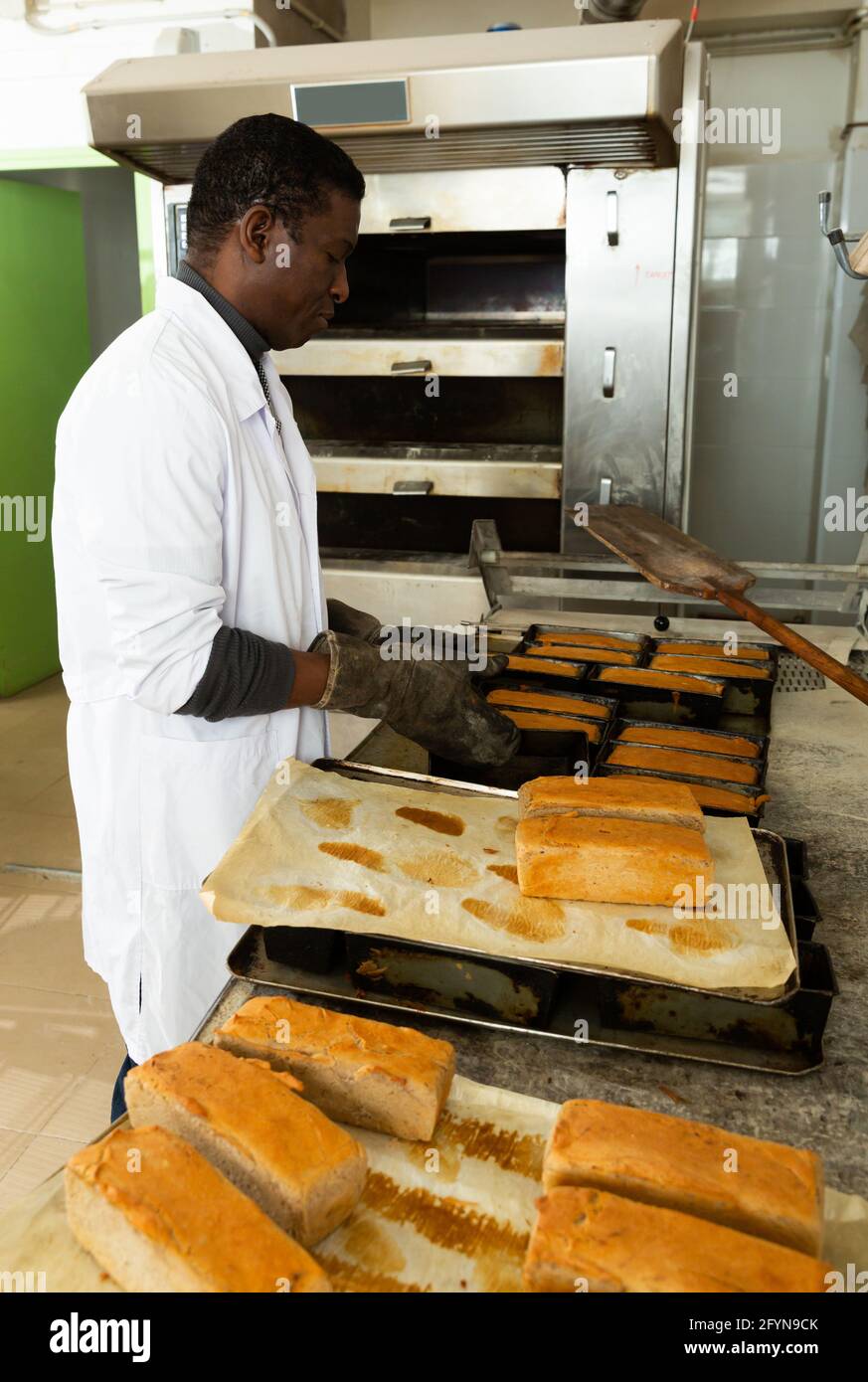 Skilled African American baker taking out freshly baked hot bread from baking molds in small bakery Stock Photo
