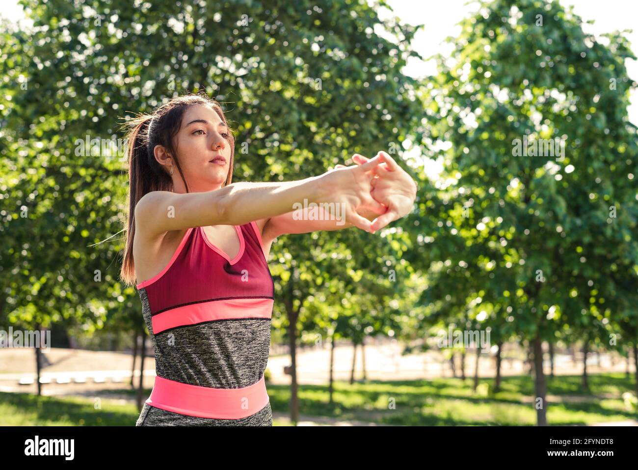 horizontal take of a beautiful woman stretching her arms. She is on a green park and wears pink and grey clothes Stock Photo