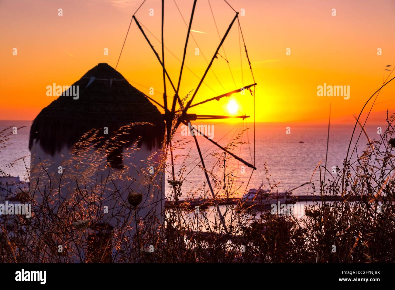 Famous traditional Greek windmill, Mykonos, Greece against sunset. Beautiful sky, sun touch sea horizon, high grass in foreground, colorful image. Stock Photo