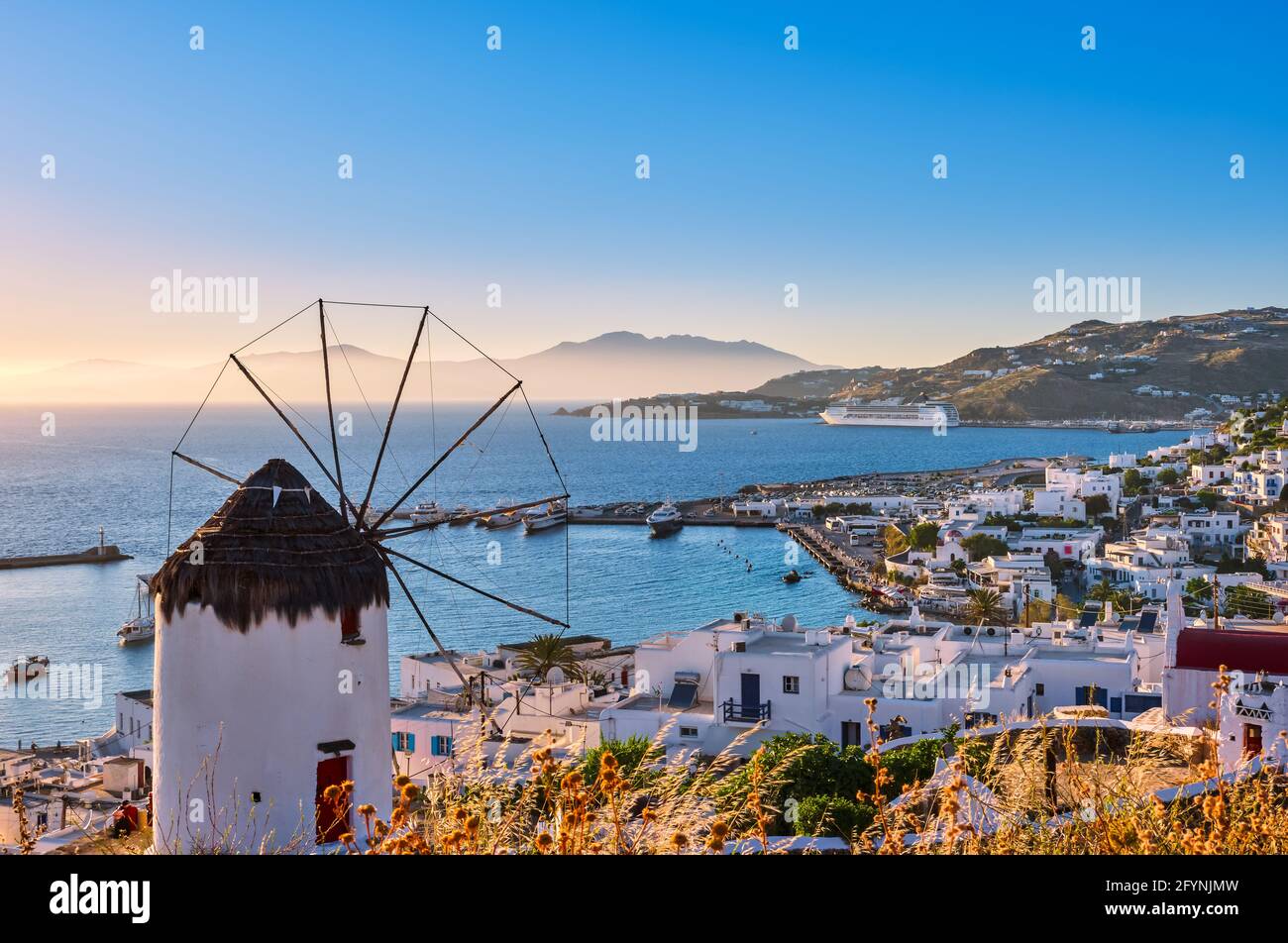 Beautiful view of Greek white windmill overlooking Mykonos, Greece at sunset. Bay, boats, cruise liner, whitewashed houses, islands, hazy background. Stock Photo