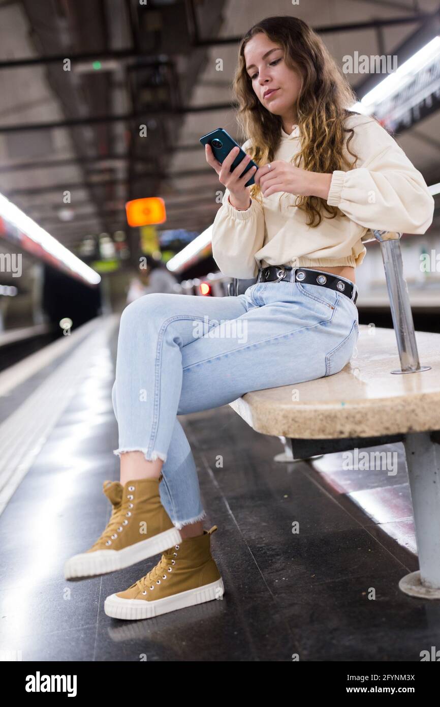 Portrait of a focused girl sitting on a subway platform bench, texing with friends on a mobile phone Stock Photo